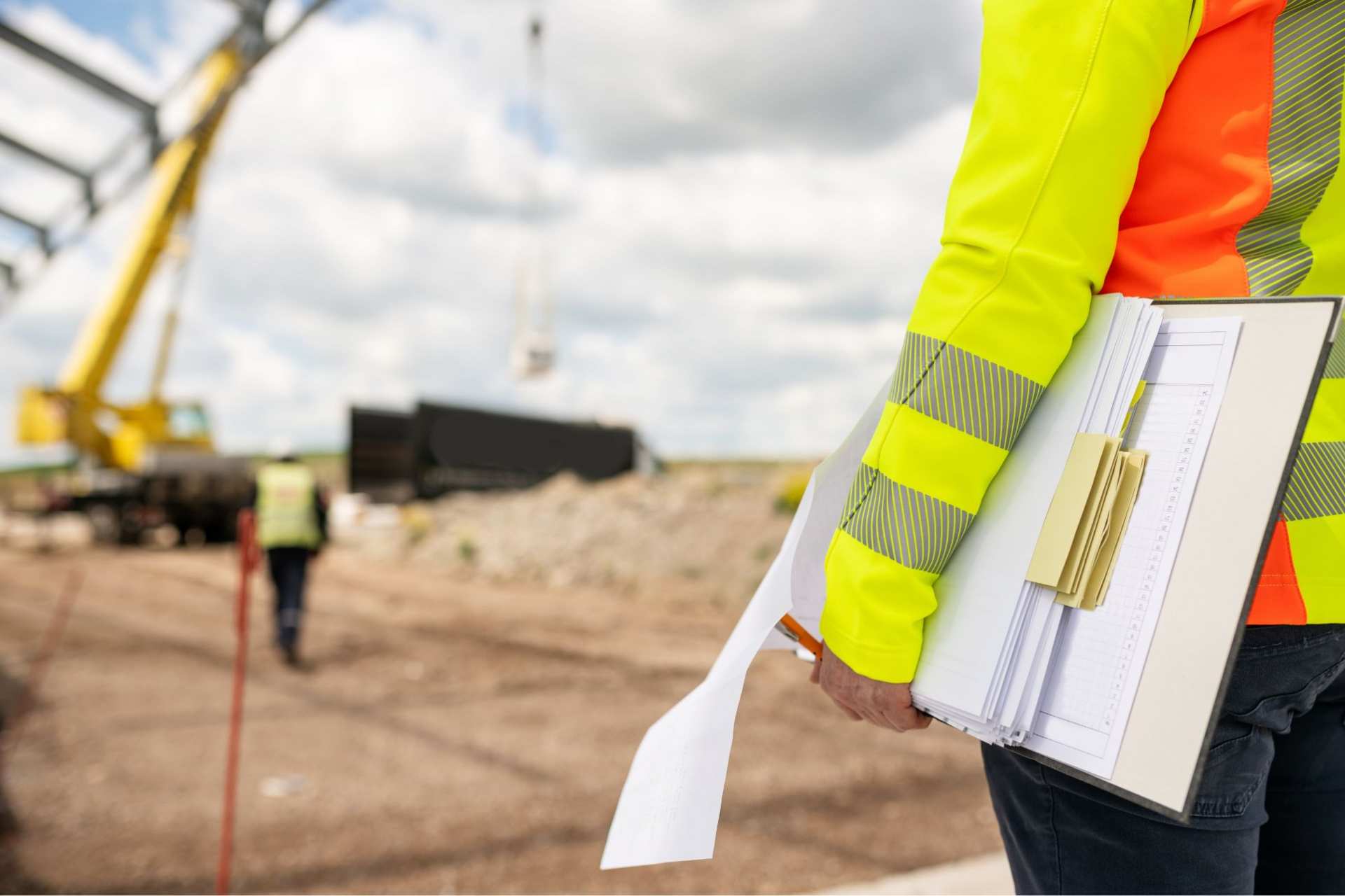Widok od tyłu na osobę w żółtej kamizelce, która trzyma plik dokumentów. W tle plac budowy.