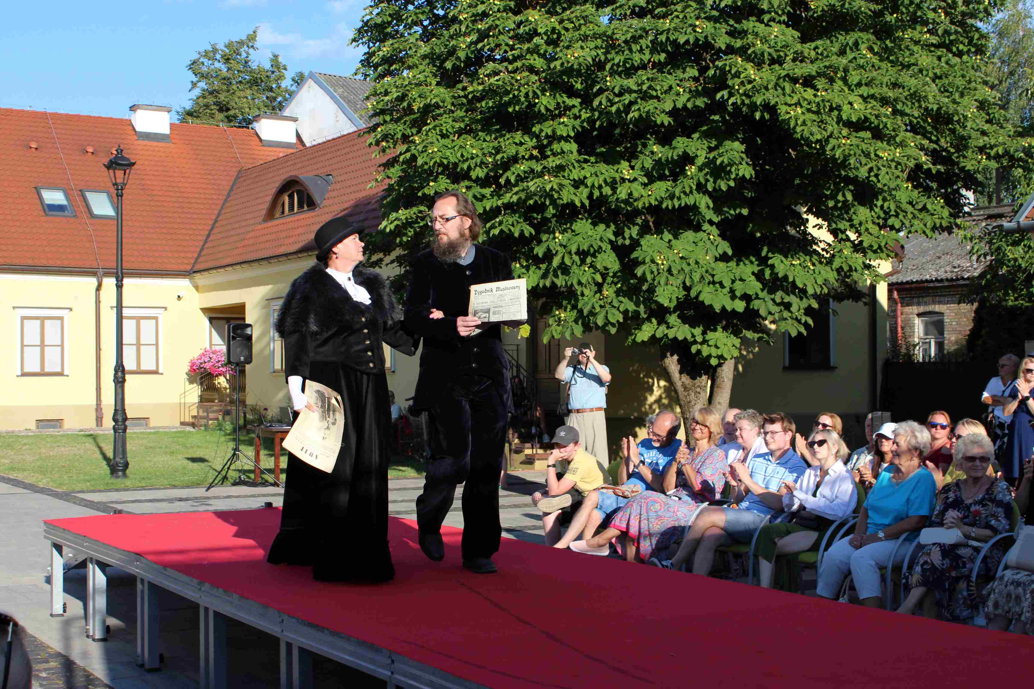 Kobieta i mężczyzna w czarnych strojach idą po podeście przykrytym czerwoną wykładziną. Obok siedzą osoby, które robią zdjęcia.
