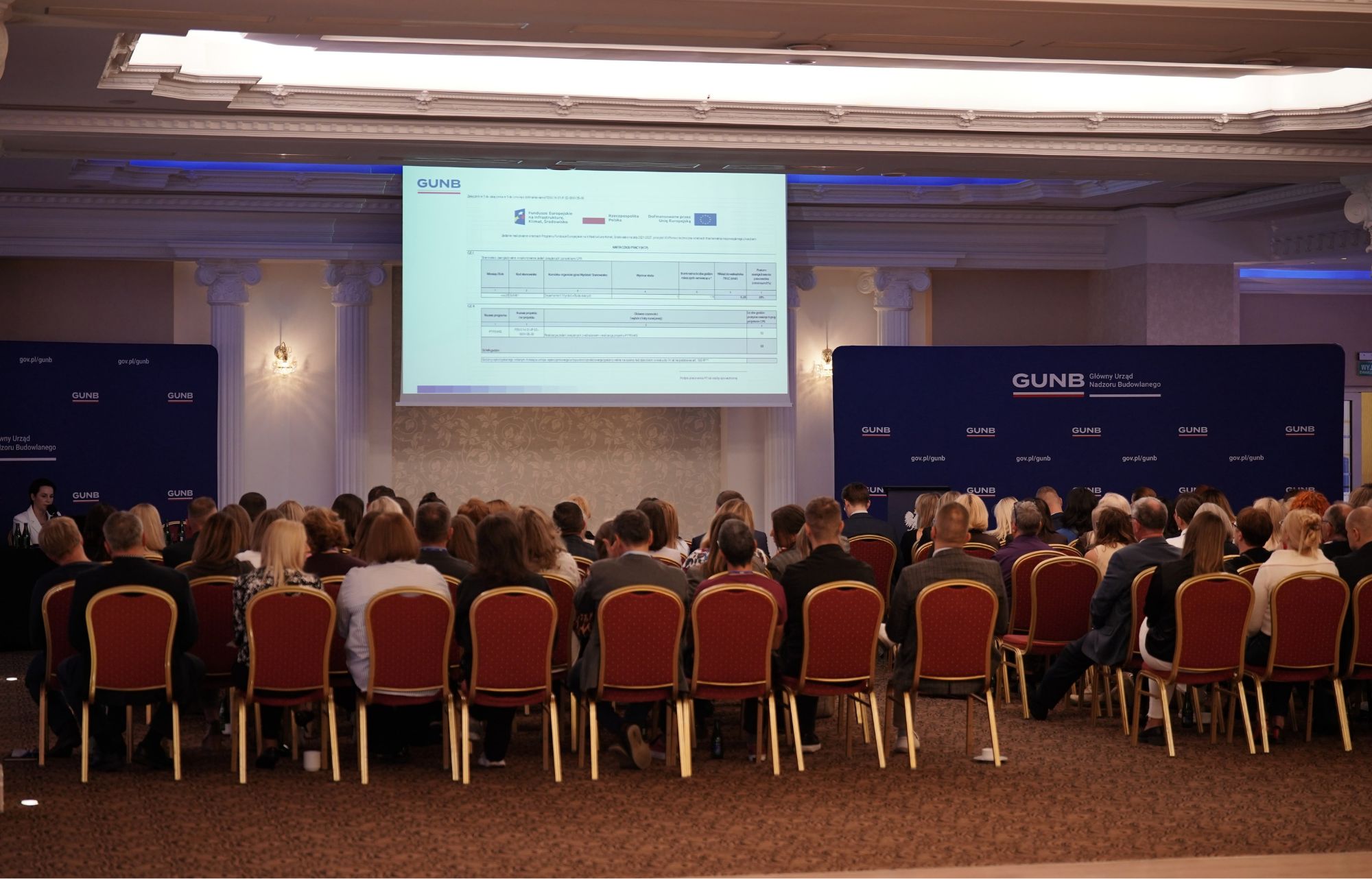 Na zdjęciu uczestnicy spotkania siedzą na krzesłach i patrzą w kierunku ekranu z prezentacją.